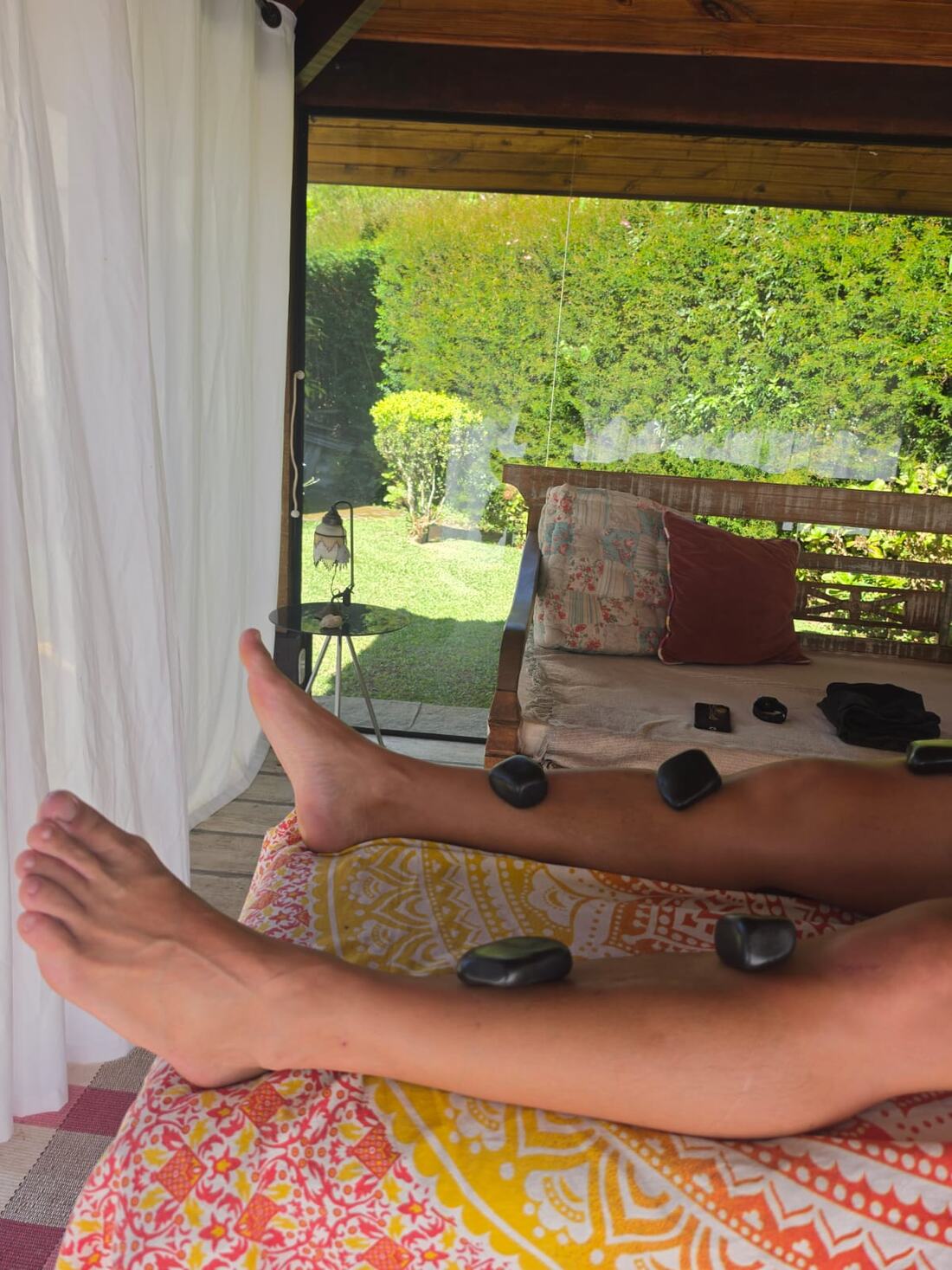 Pernas apoiadas com pedras quentes vulcânicas durante sessão de massagem, em varanda com vista para o jardim